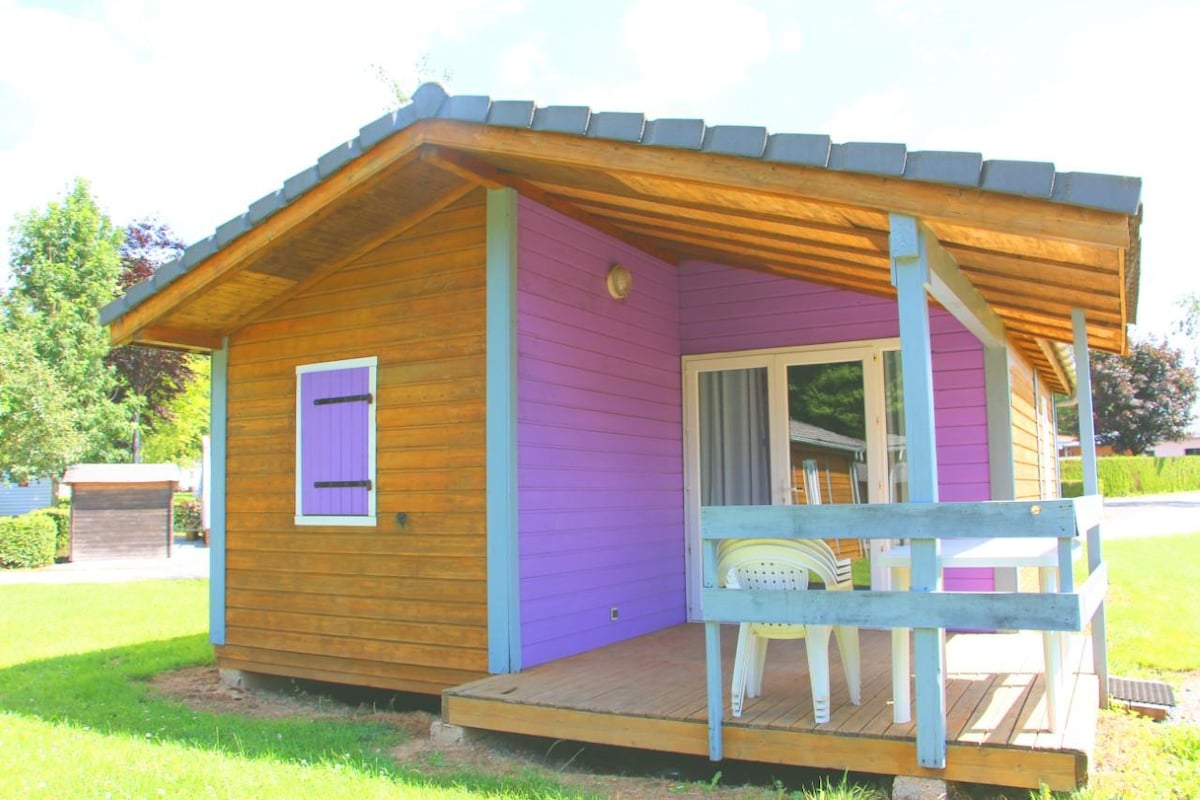 A wooden chalet with a colorful purple and natural wood exterior stands in a grassy area. A covered terrace is equipped with a white table and chairs. Large windows reflect sunlight, providing a bright and airy atmosphere within.