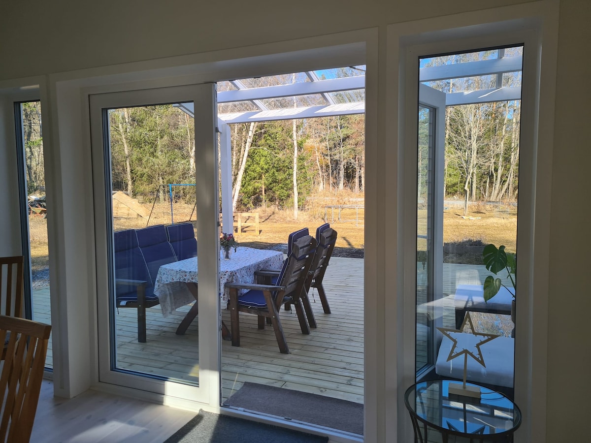 Large glass doors open to a sunlit terrace featuring a wooden deck. Outdoor seating is arranged around a table with a tablecloth, surrounded by trees and open space. A small table with a decorative star is positioned indoors, offering a view of the outdoor area.