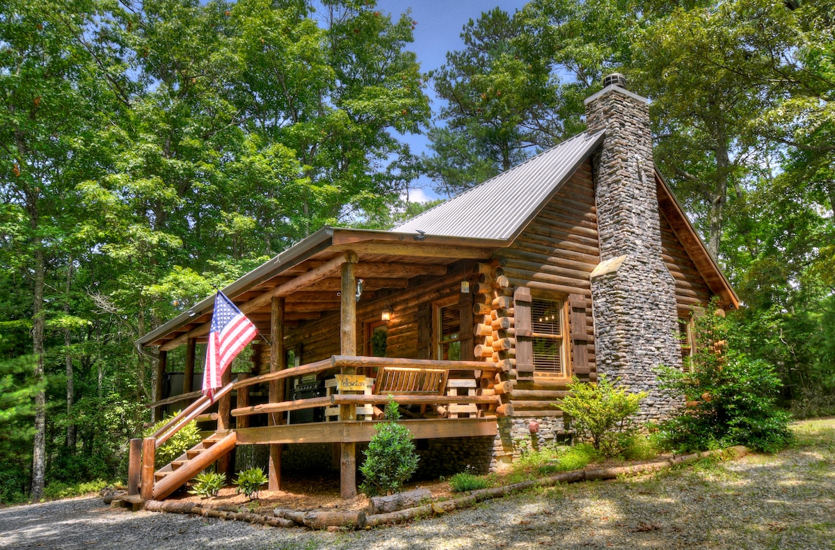 A charming log cabin is framed by lush greenery, showcasing a sturdy stone chimney and a welcoming porch. An American flag gently flutters beside the entrance, which features wooden steps and surrounding landscaped areas. Natural light fills the space, highlighting the cabin's rustic design.