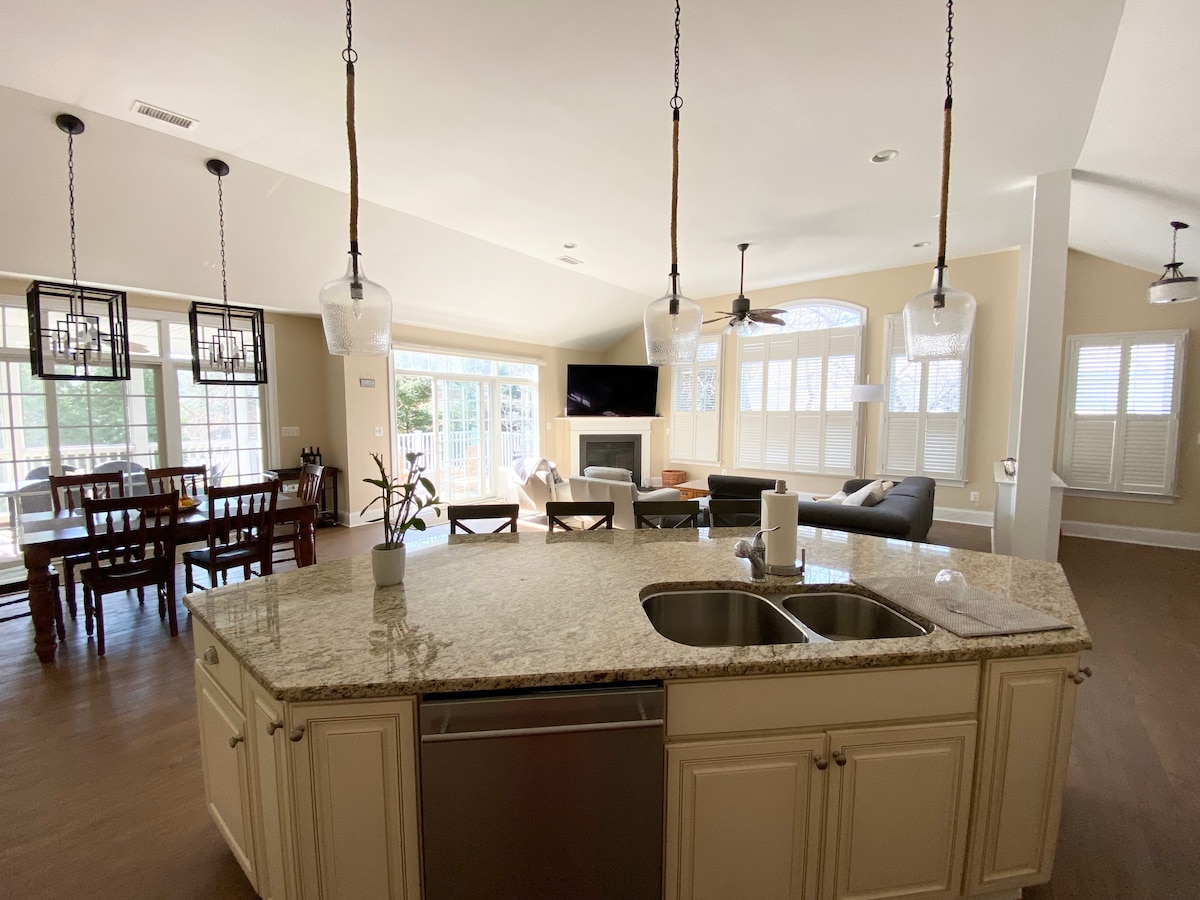 An open concept living area is observed, featuring a spacious kitchen island with seating and granite countertops. Natural light floods the room through large windows, illuminating the dining area with a wooden table and chairs. The seating area includes a modern sofa and a fireplace.