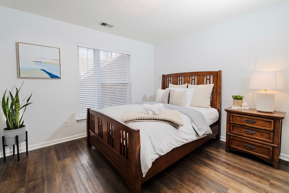 A bright bedroom features a spacious wooden bed with crisp white linens and multiple pillows. A nightstand is positioned beside the bed, accentuated by a lamp. Natural light enters through a partially covered window, and a potted plant adds a touch of greenery.