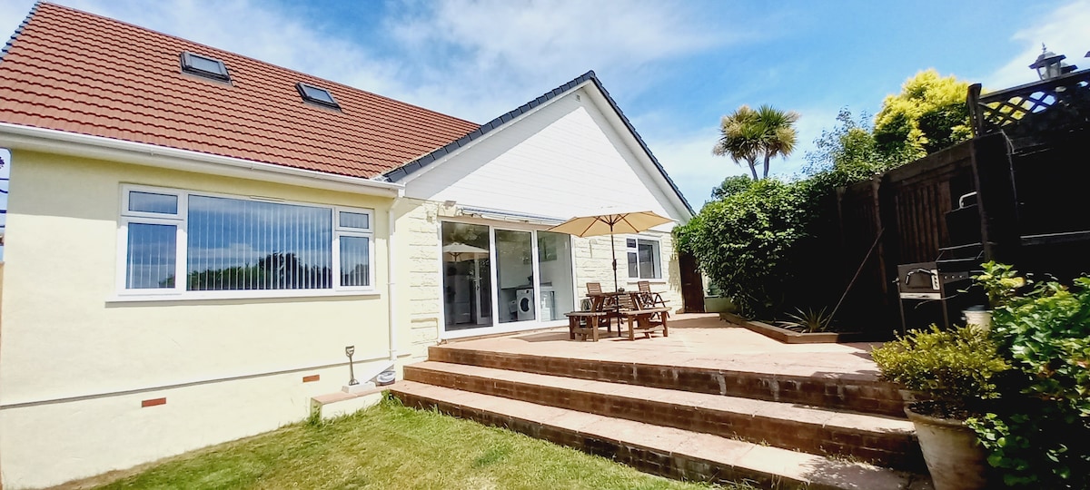 The exterior of the home showcases a spacious patio area with wooden furniture under a large umbrella, providing a relaxing space for outdoor meals. Elevated steps lead up to the entrance, framed by lush greenery and an inviting garden.