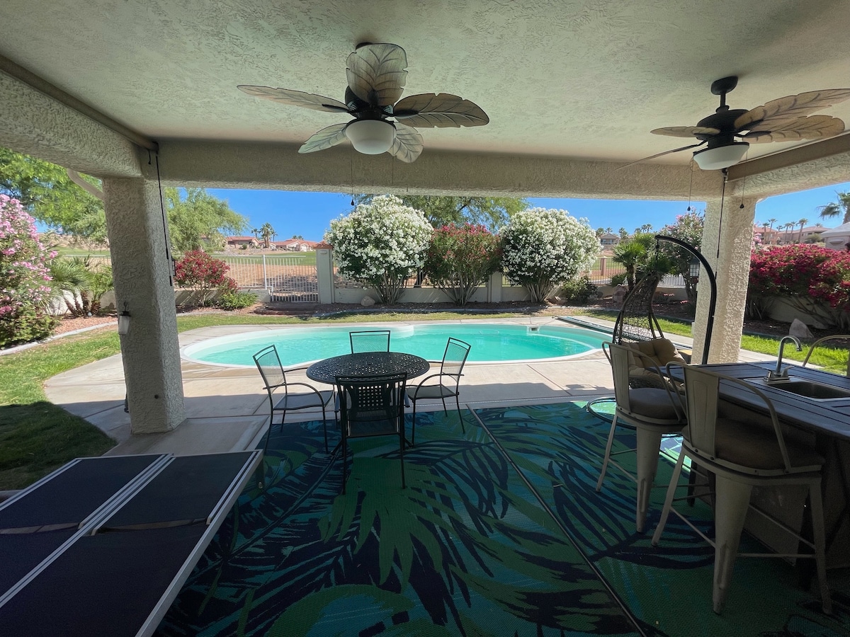 A covered patio area is featured, overlooking a private pool surrounded by lush greenery. Comfortable seating is provided by a round table and chairs, with light streaming in through the outdoor space, creating a serene environment for relaxation.