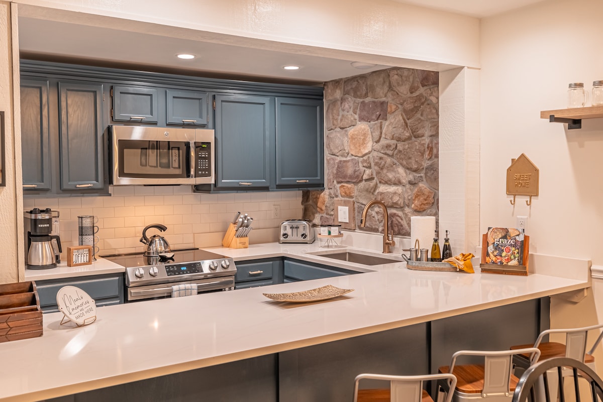 The kitchen features modern appliances including a stainless steel microwave and stove. Blue cabinets are complemented by a stone accent wall. A bar-style countertop with seating faces an open space, creating an inviting area for meal preparation and enjoyment.