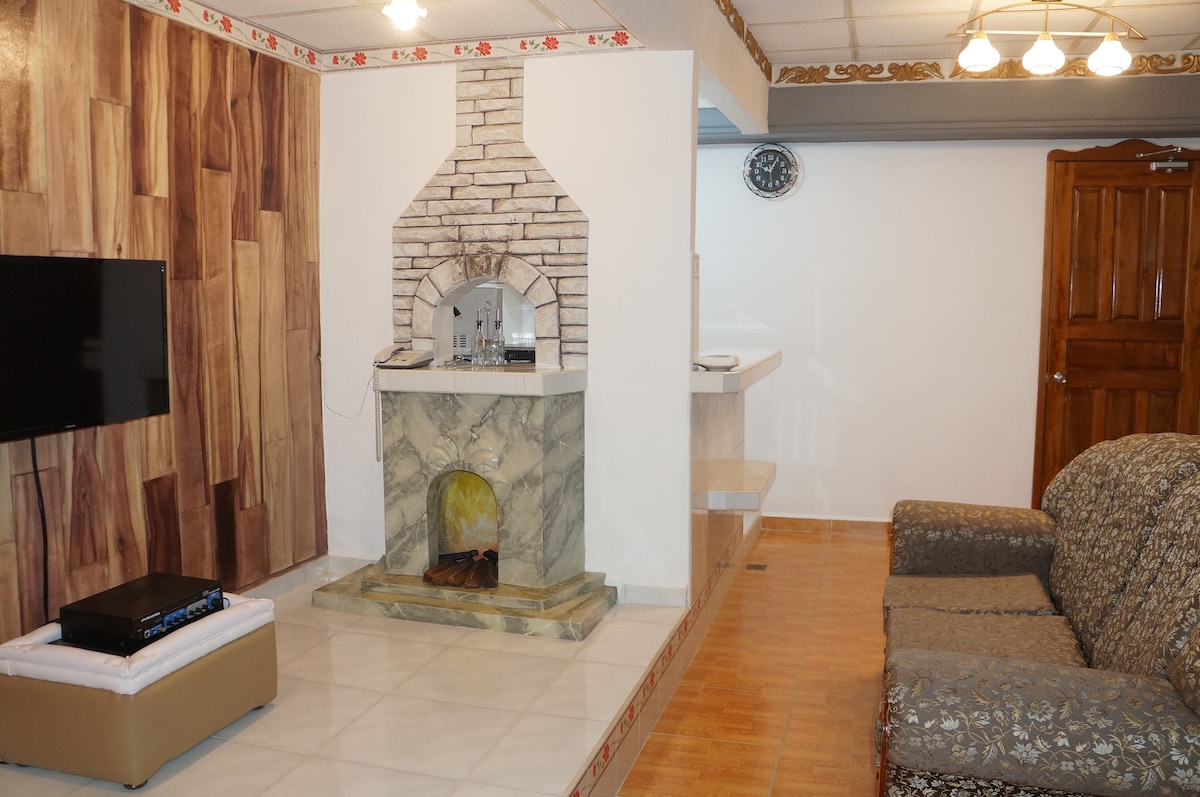 The living area features a stone fireplace framed by a light-colored wall. A flat-screen television is mounted on one wall, and a comfortable couch is positioned nearby. Wooden elements are visible, enhancing the warm ambiance of the space.