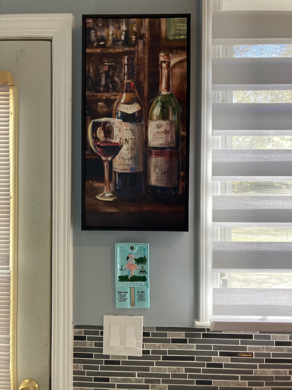 A framed painting of wine bottles and a glass of red wine is displayed against a gray wall. Below, a decorative wall plaque featuring a woman is attached to the wall near light switches. The kitchen area showcases a stylish and welcoming atmosphere.