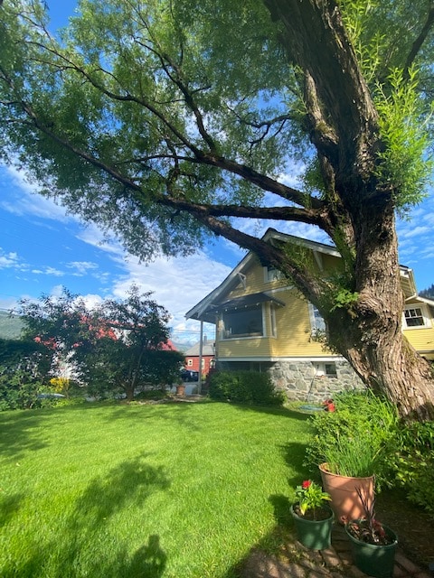 A well-maintained outdoor space features a lush green lawn surrounded by colorful flower pots. A large tree provides shade, and a charming yellow house with large windows is partially visible in the background against a clear blue sky.