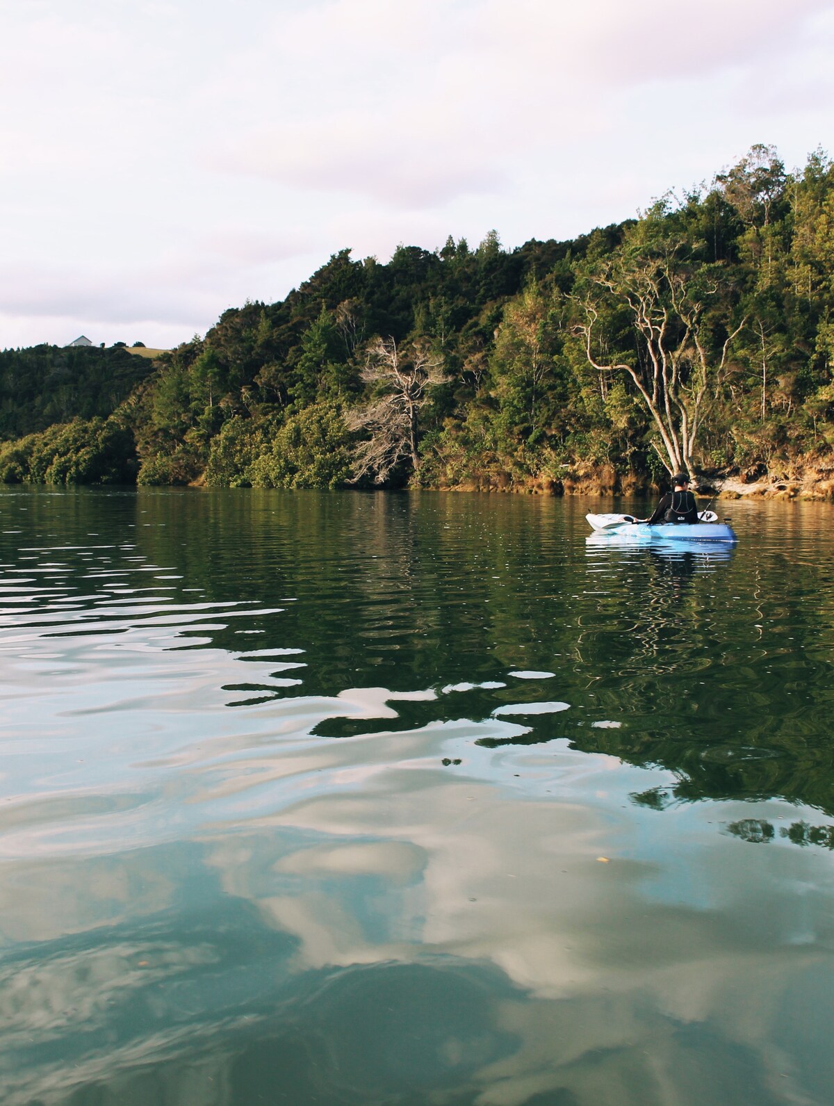 Calm waters reflect the surrounding verdant hills and trees along the river. A kayak is seen in the foreground, gliding smoothly on the serene surface. The backdrop features lush green foliage, creating a tranquil atmosphere.