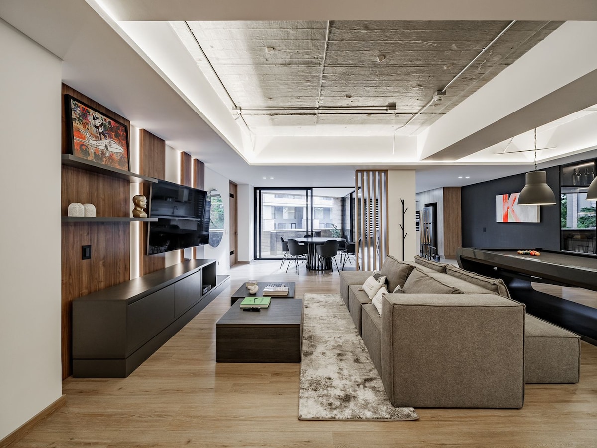 A spacious living area features modern furnishings and an open layout, with a large sectional sofa facing a sleek entertainment unit. Soft lighting highlights the neutral color palette and natural materials. A dining area is visible in the background, showcasing a contemporary table and chairs.
