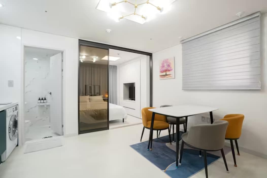 A modern living area is present, featuring a round dining table with four chairs, two of which are upholstered. A washing machine is located near the entrance, while the bedroom area is separated by sliding glass doors, showcasing a cozy sleeping space in the background.