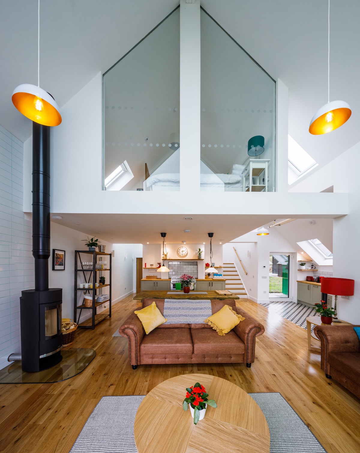 An open-plan living area is highlighted by high ceilings and large windows allowing natural light. A cozy seating arrangement with a brown sofa faces a central wooden table, while a sleek wood stove adds warmth. A staircase leads to a loft space visible above.