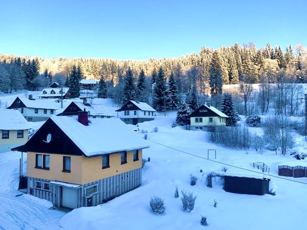 A quiet winter landscape features several chalets nestled among snow-covered trees. The sun casts a soft glow over the scene, illuminating the rooftops and surrounding white terrain. Snow blankets the ground, enhancing the serene atmosphere of the Kořenov area.