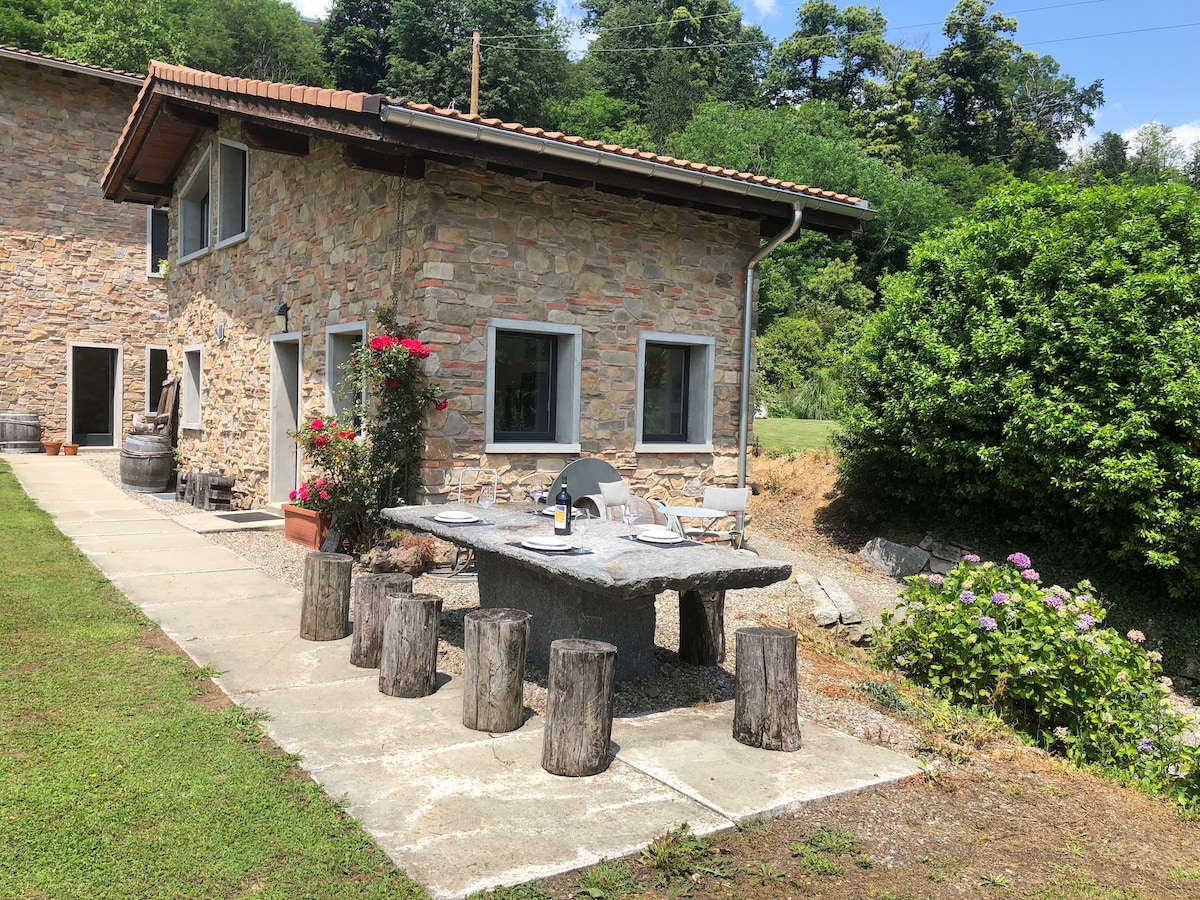 An outdoor dining area features a large granite table surrounded by rustic wooden stools. The stone building shows large windows and a welcoming entrance. Lush greenery and blooming flowers enhance the natural setting. A smooth path leads to the dining space from the property.
