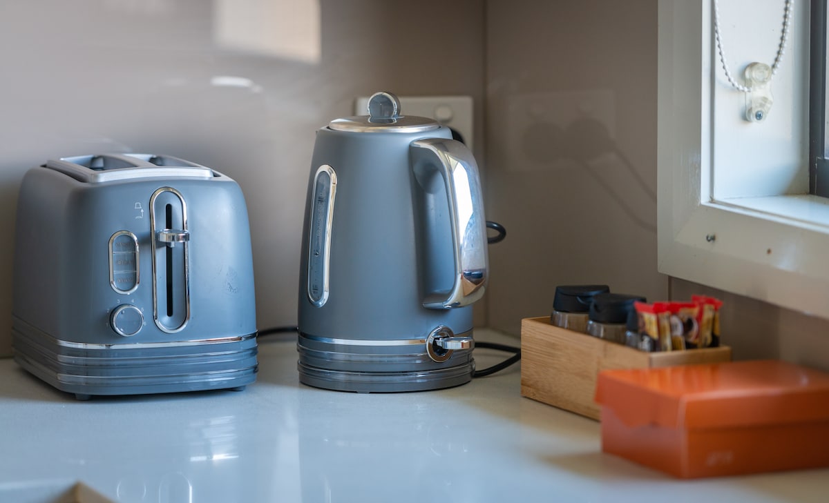 A modern kitchen countertop is displayed, featuring a silver toaster and kettle. A wooden container holds various tea and coffee supplies, accompanied by an orange box. The setting is well-lit, with a clean and organized appearance.