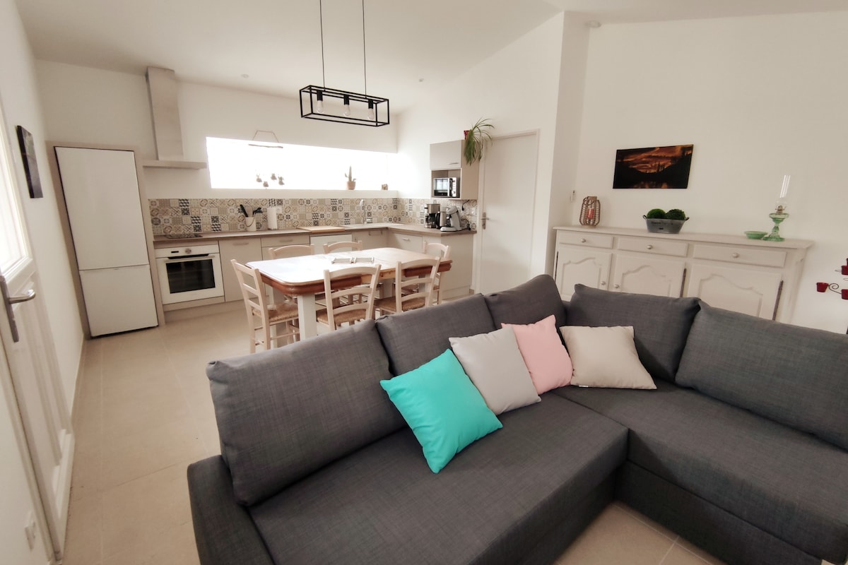 A modern living area features a gray sectional sofa with colorful accent pillows. An open kitchen with tiled backsplash is visible, showcasing wooden dining furniture and ample natural light from the window. Clean lines and neutral colors create a welcoming space.