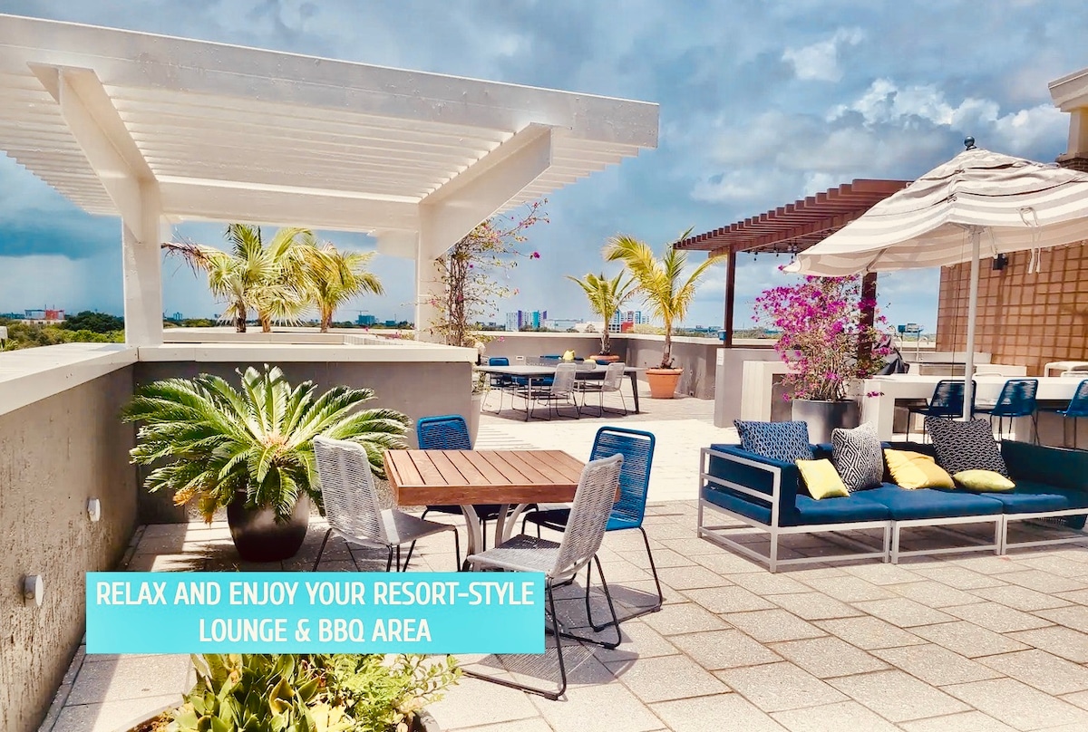 A spacious rooftop lounge area is shown, featuring a mix of seating options, including lounge chairs and dining tables. Lush greenery complements the setting, where vibrant flowers add color. A shaded area is provided by a pergola, enhancing comfort for outdoor relaxation.