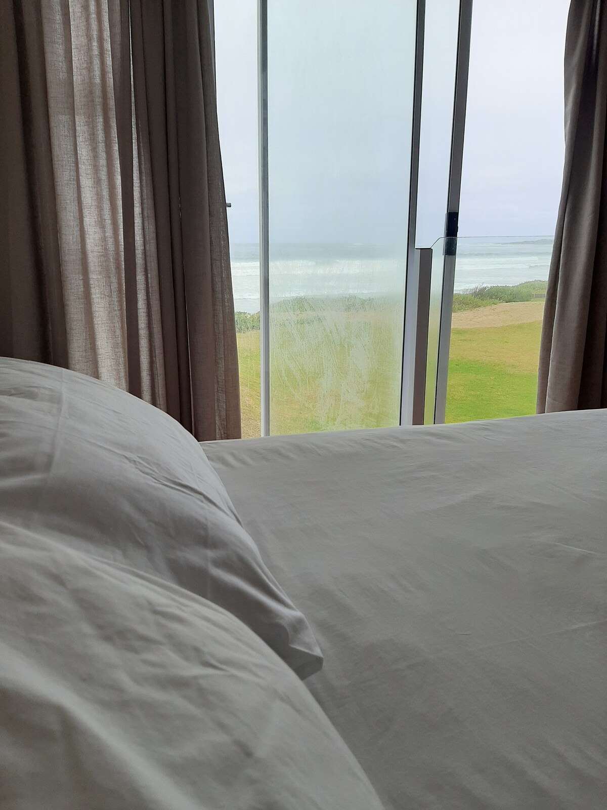 A spacious double bed with crisp white linens is positioned near a large glass window. Light sheer curtains frame the window, which offers an uninterrupted view of the ocean and grassy landscape beyond.