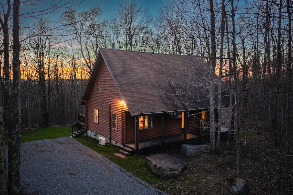 A charming log cabin is set amidst a wooded landscape. Warm lights illuminate the exterior, providing a welcoming glow as twilight descends. The gravel driveway leads up to the front porch, surrounded by natural scenery and a sense of tranquility.