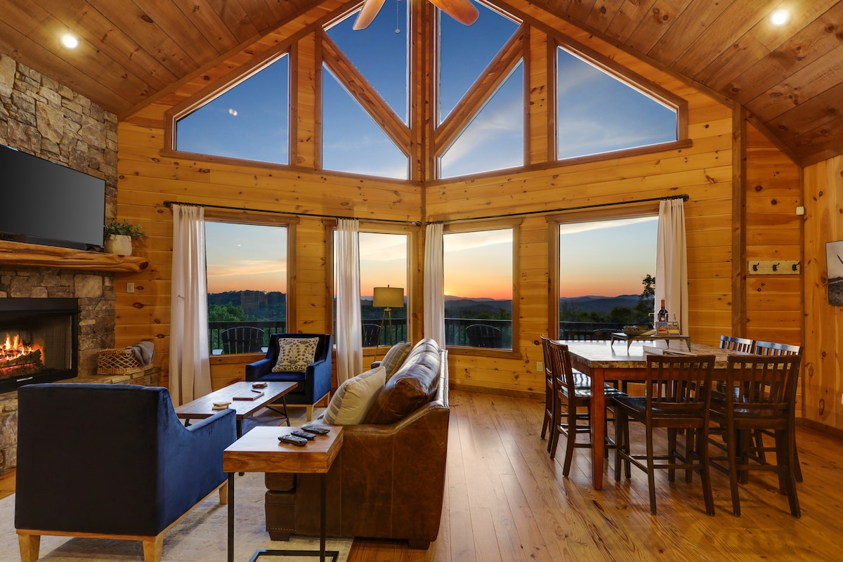 A spacious living area features a stacked stone gas fireplace, large leather sofa, and two side chairs. Floor-to-ceiling windows provide expansive views of the mountains and sunset. A wooden dining table with multiple chairs is positioned nearby, enhancing the open and inviting atmosphere.