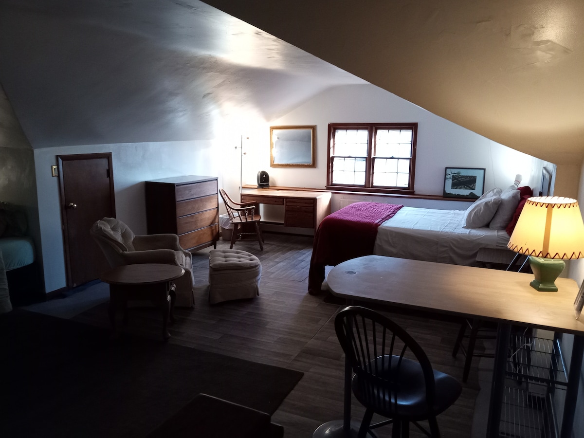 A spacious room features a double bed, a seating area with two armchairs, and a small round table. Natural light enters through two windows, enhancing the neutral-toned decor. A dresser and desk provide functional space, while a soft lamp adds warmth.