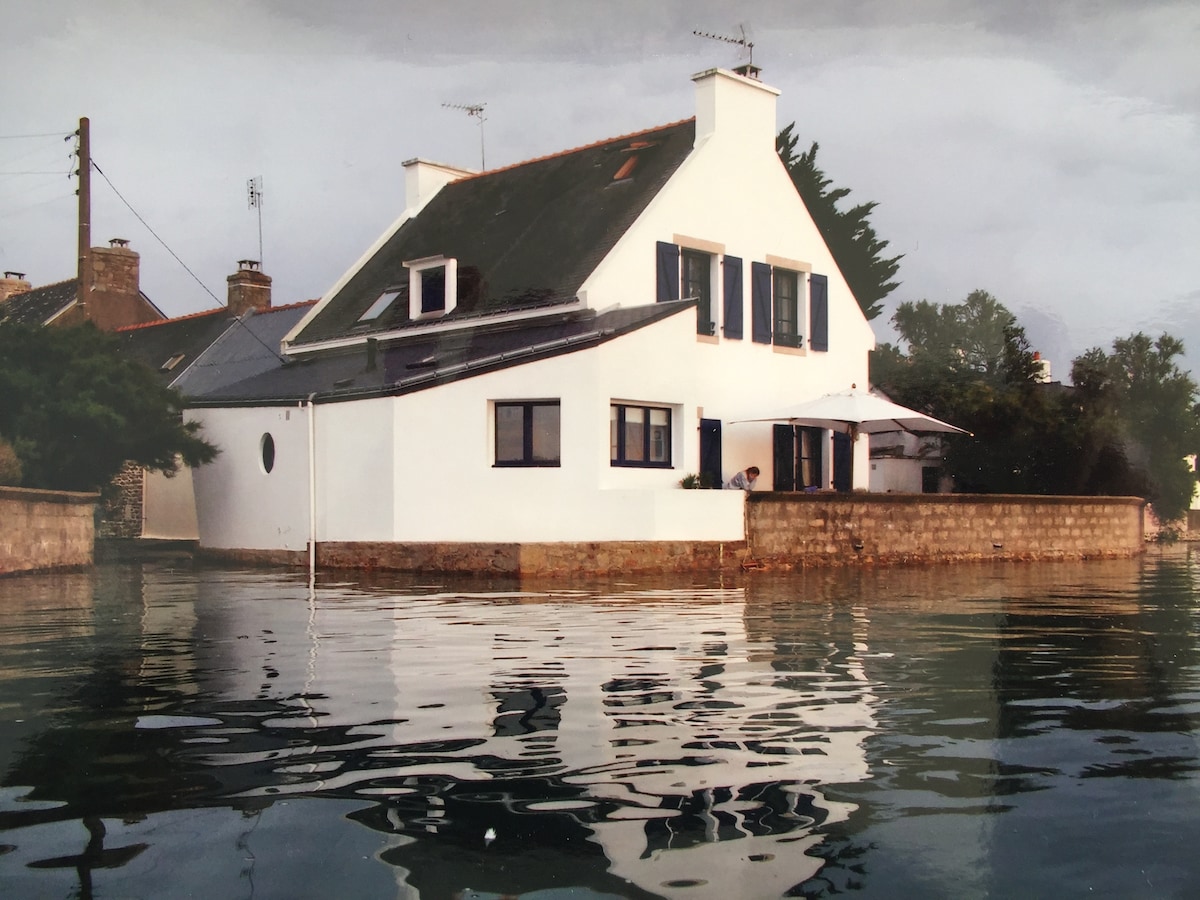 A modern two-story house is depicted, featuring a white exterior and dark shutters. The home is set beside calm water, reflecting its structure beautifully. Lush greenery surrounds the property, and a small terrace can be seen, suggesting a welcoming space for relaxation.
