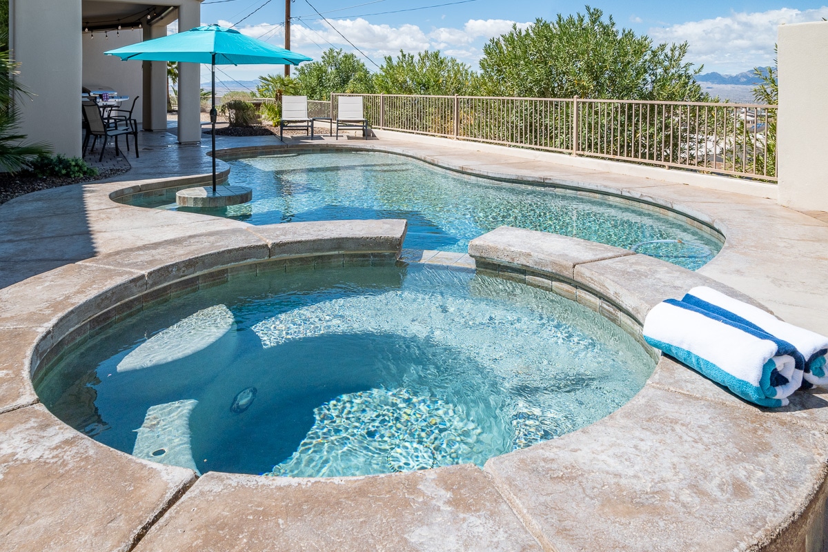 A large pool and hot tub area are visible, with clear water reflecting the surroundings. A light blue umbrella provides shade over the seating area, while a stack of white towels with blue accents rests nearby. Lush greenery borders the space, enhancing the tranquil atmosphere.