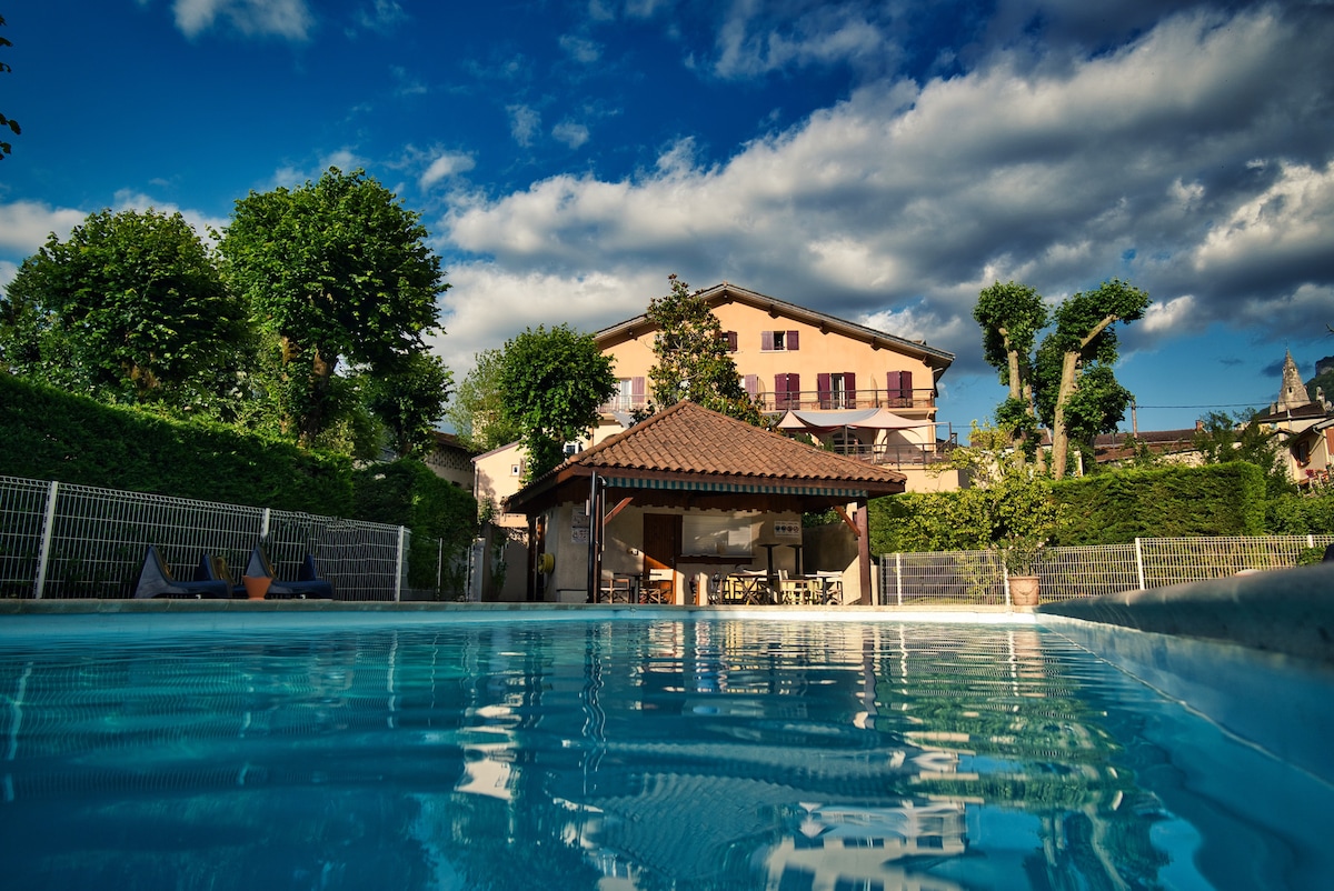 The image displays a refreshing outdoor pool framed by lush greenery. A charming building with purple windows and a tiled roof is visible in the background. An outdoor seating area under a shaded structure offers space for relaxation. A clear blue sky contrasts with the surrounding trees.