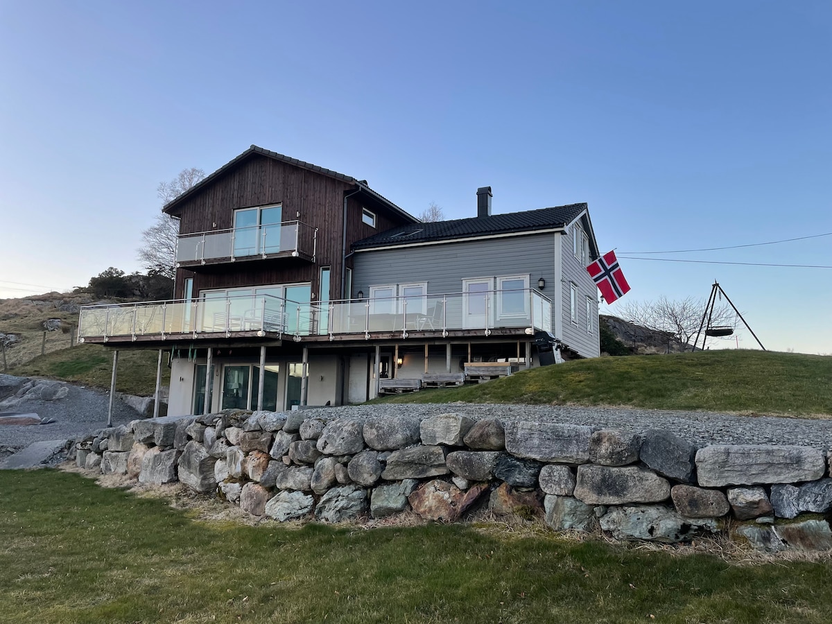 A three-story house is presented in a peaceful setting, featuring a mix of wooden and modern elements. Glass railings line the spacious deck, which overlooks a grassy area and stone wall. A Norwegian flag adorns the exterior, signaling a welcoming atmosphere.