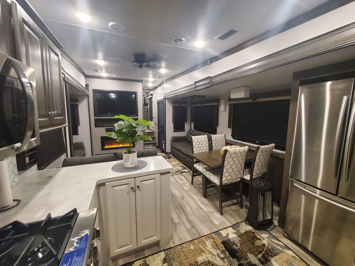 An inviting interior of the RV showcases a modern kitchen with a marble countertop and stainless-steel appliances. A cozy dining table sits next to comfortable seating, while a sleek fireplace and large windows enhance the open layout. Soft lighting adds warmth to the space.
