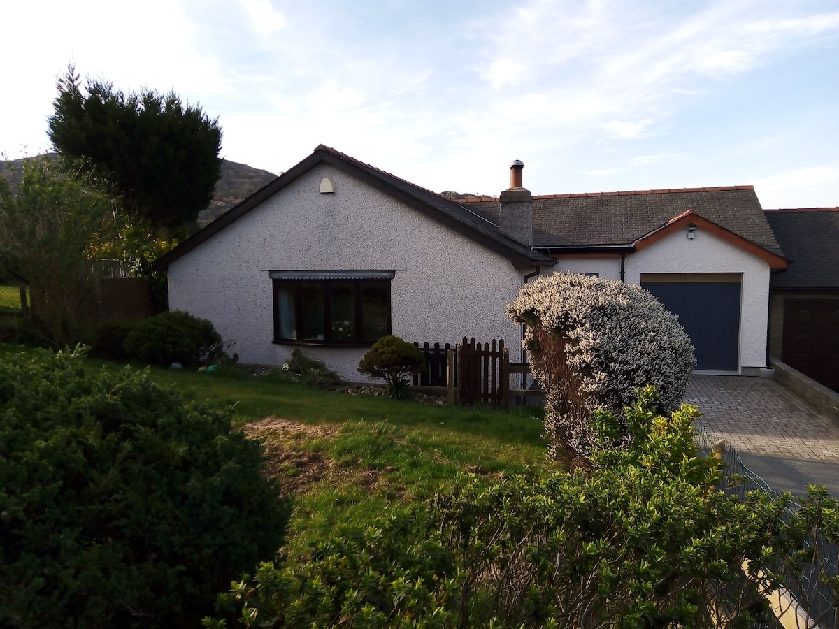 The exterior of a single-story bungalow is displayed, featuring a neatly maintained front garden with shrubs and grass. A paved driveway is visible, leading to a garage on the right side. Large windows offer a glimpse into the living area, complemented by a sloped roof.