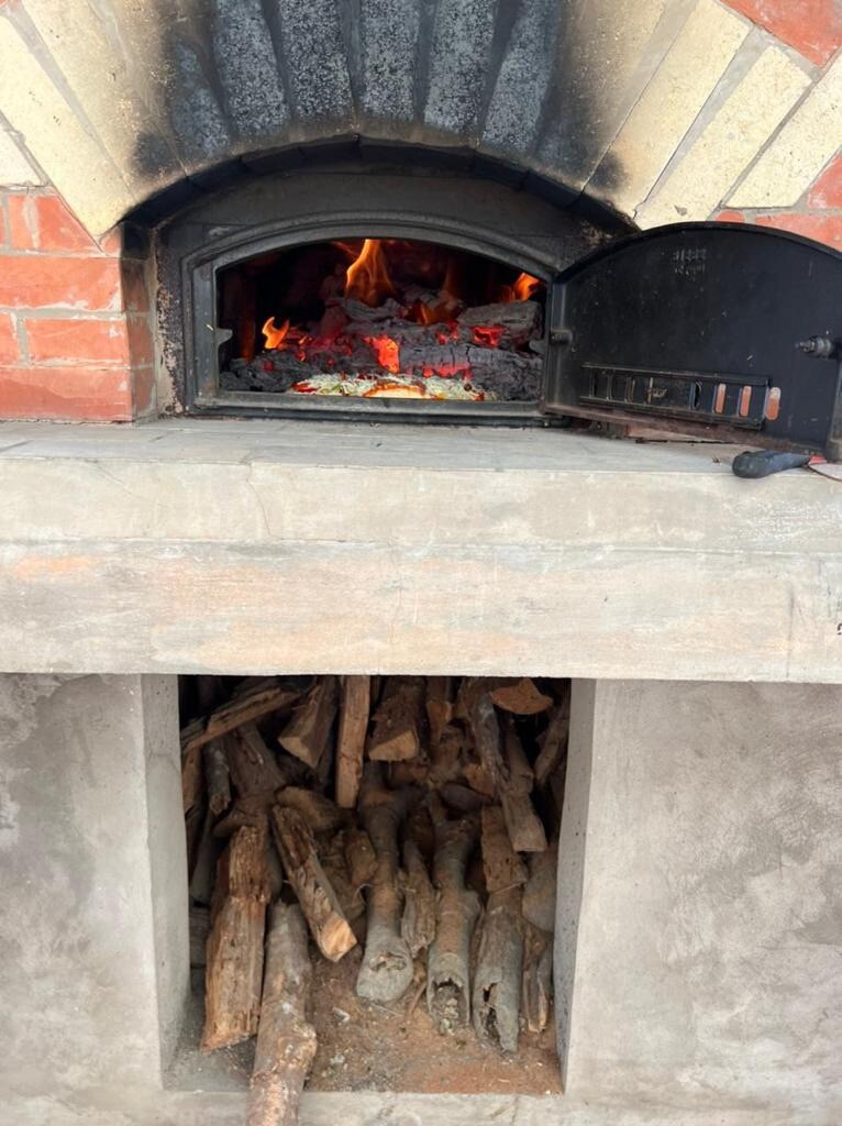 An outdoor brick oven displays a glowing ember-filled interior, with flames visible among the ashes. Below, a neatly organized storage area holds an assortment of firewood, ready for use in cooking. The structure is designed for preparing bread and pizza.