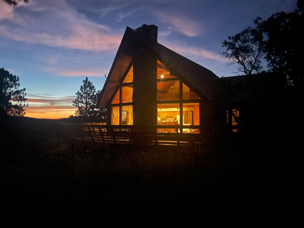 The mountain home is illuminated against a twilight sky, showcasing wooden architectural details. A spacious deck with seating is visible, creating a seamless connection with nature. Warm light glows from large windows, highlighting the inviting interior and expansive kitchen space.