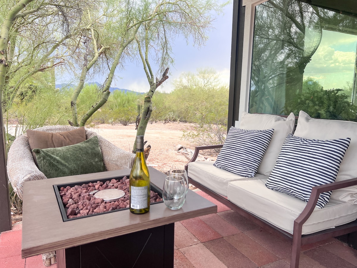 An outdoor seating area is shown, featuring a cozy couch with three striped pillows and a comfortable armchair. A fire pit table is positioned in front, adorned with decorative stones. The surrounding landscape includes desert vegetation, providing a serene atmosphere.