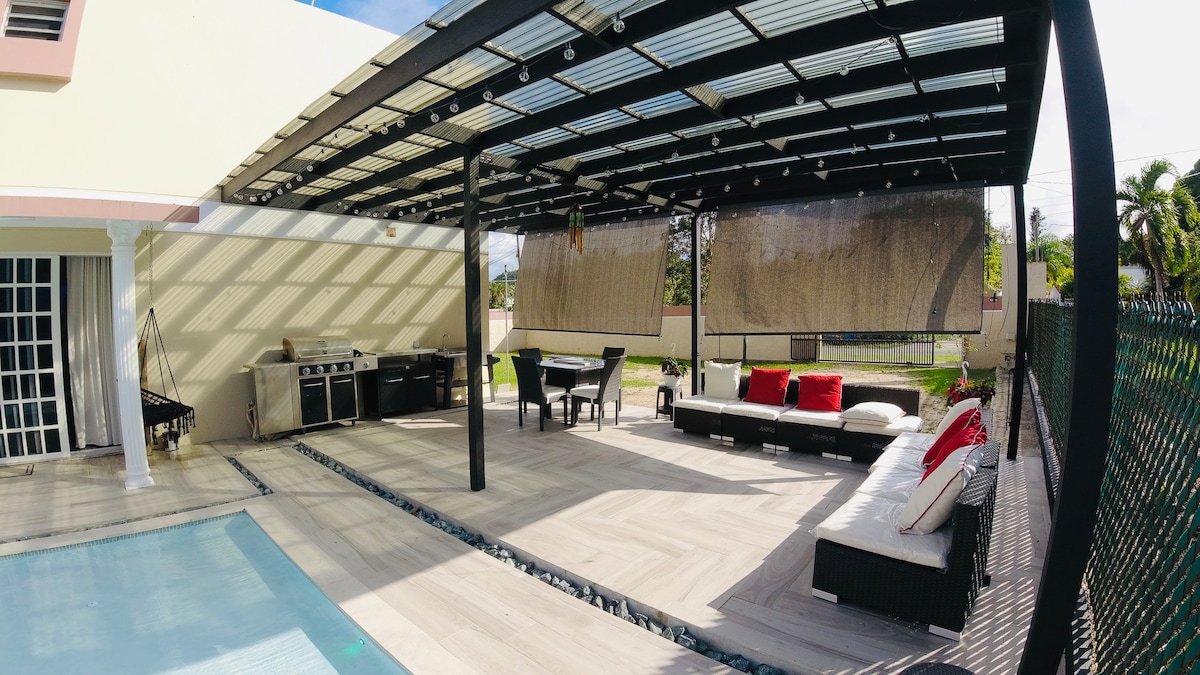 An outdoor lounging area features modern furniture arranged beneath a pergola with a transparent roof. A dining table is complemented by a grill station, while a tan sectional couch adorned with red cushions provides a comfortable seating option. Natural light creates a playful interplay of shadows on the floor.