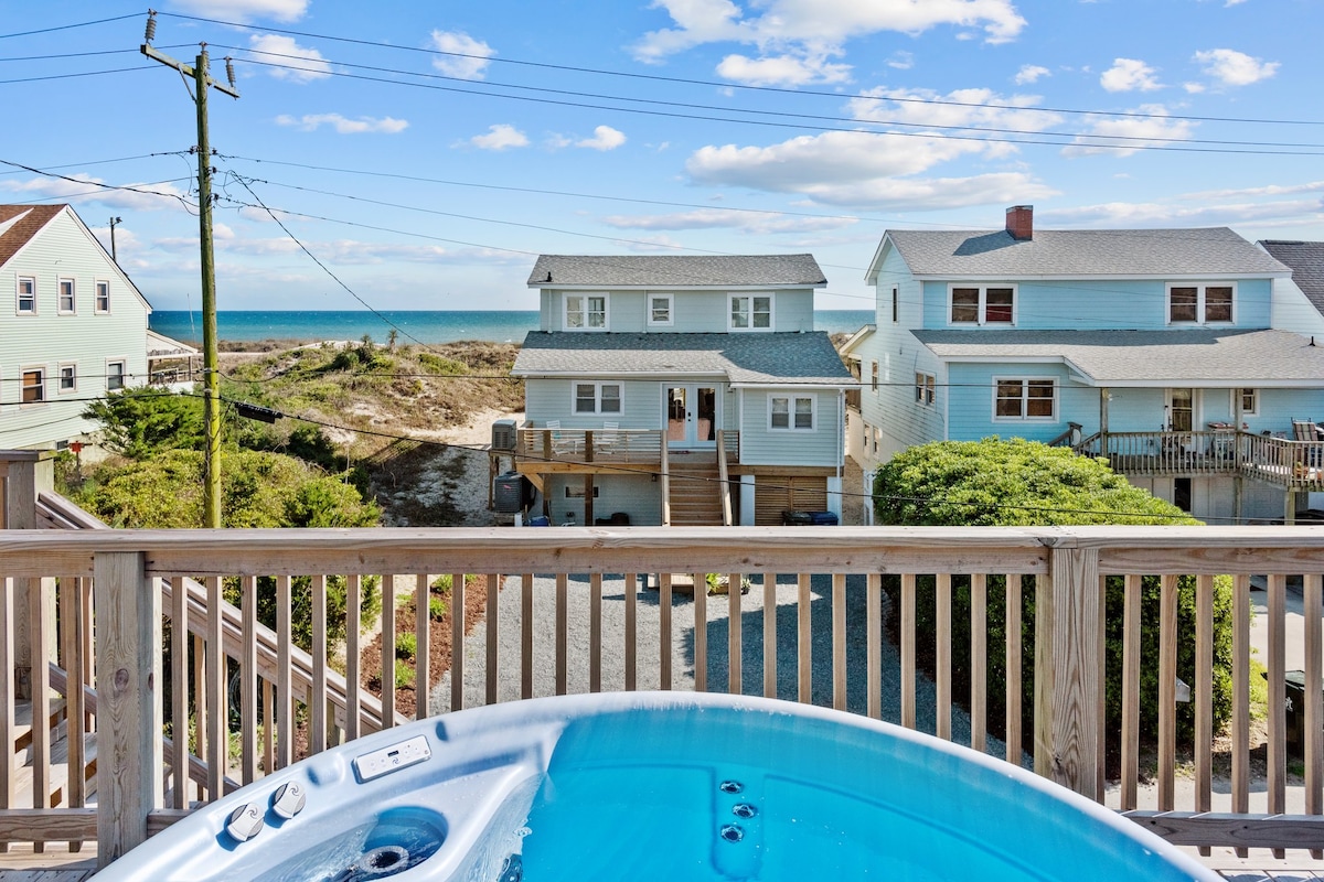 A view of the ocean from a private second-story hot tub, showcasing the blue water and sky. Nearby houses are visible among lush greenery, and the outdoor deck railing frames the scene, offering a serene atmosphere for relaxation.