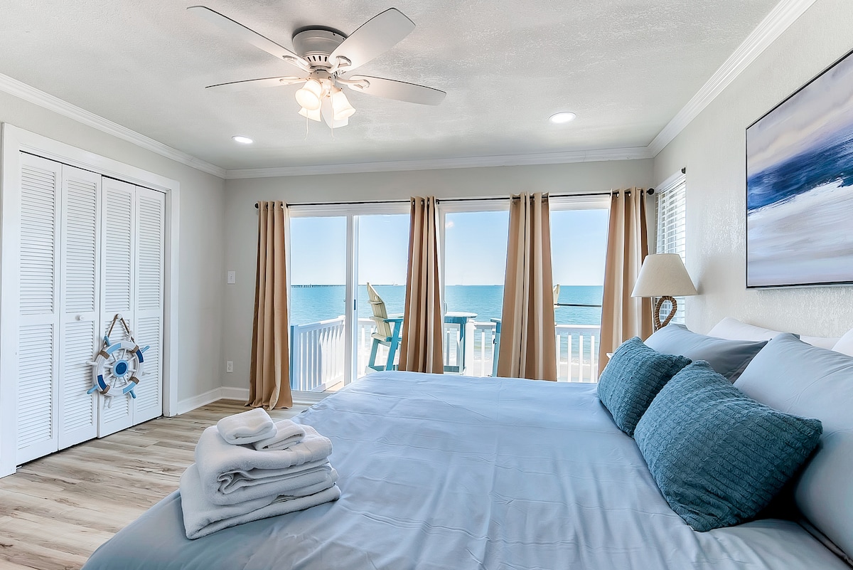 A serene master bedroom features a spacious bed with light blue bedding and accented with matching pillows. Large windows, dressed in soft curtains, offer a sweeping view of the ocean and access to a private deck. A ceiling fan provides additional comfort.
