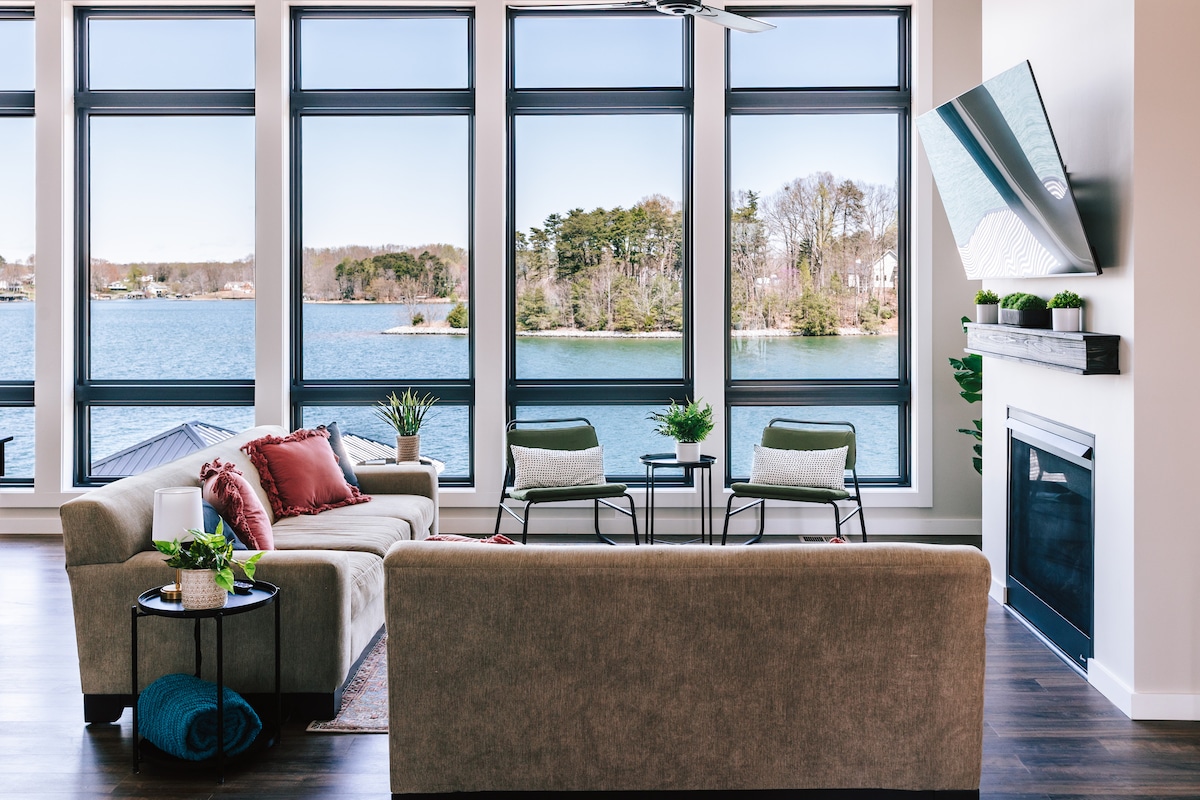 A modern living area features large windows that provide a view of the lake, allowing natural light to illuminate the space. A comfortable sofa and two chairs are arranged around a coffee table, with a fireplace and a television mounted above a stylish mantle.