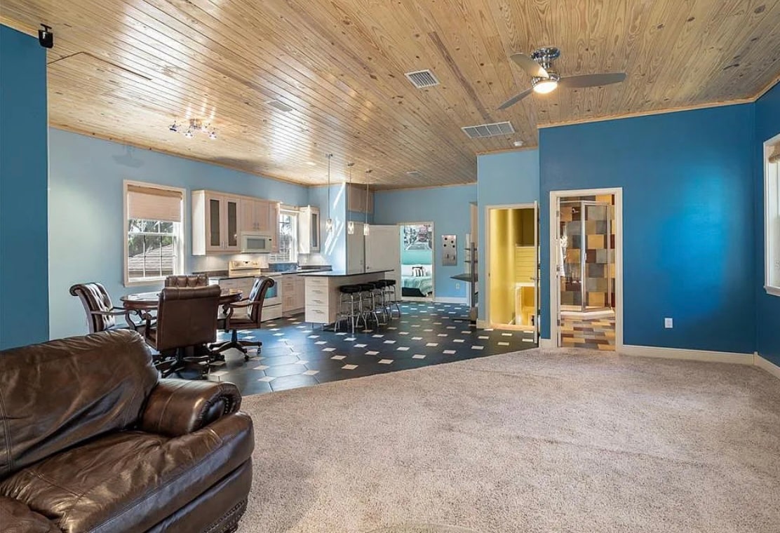 An open living space features a cozy seating area with a dark brown couch and a ceiling fan above. A full kitchen with modern appliances is visible, along with a dining area containing a wooden table and leather chairs. Colorful tiles add vibrancy to the flooring.