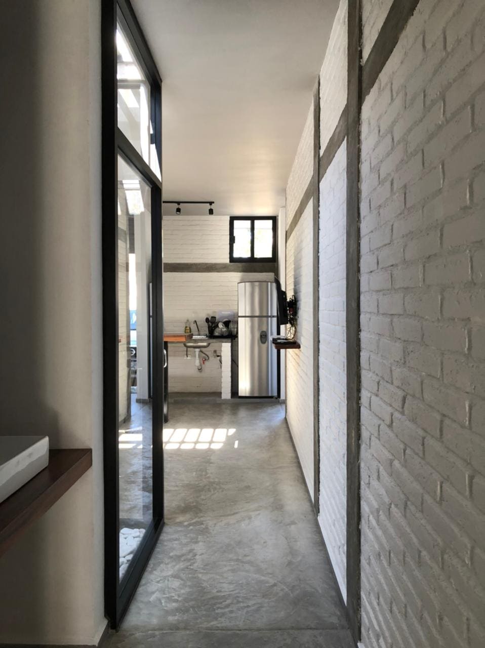 A sleek, modern kitchen is accessible through a long corridor featuring white brick walls. Large windows allow natural light to illuminate the space, highlighting the stainless steel refrigerator and minimalist cabinetry along the wall.