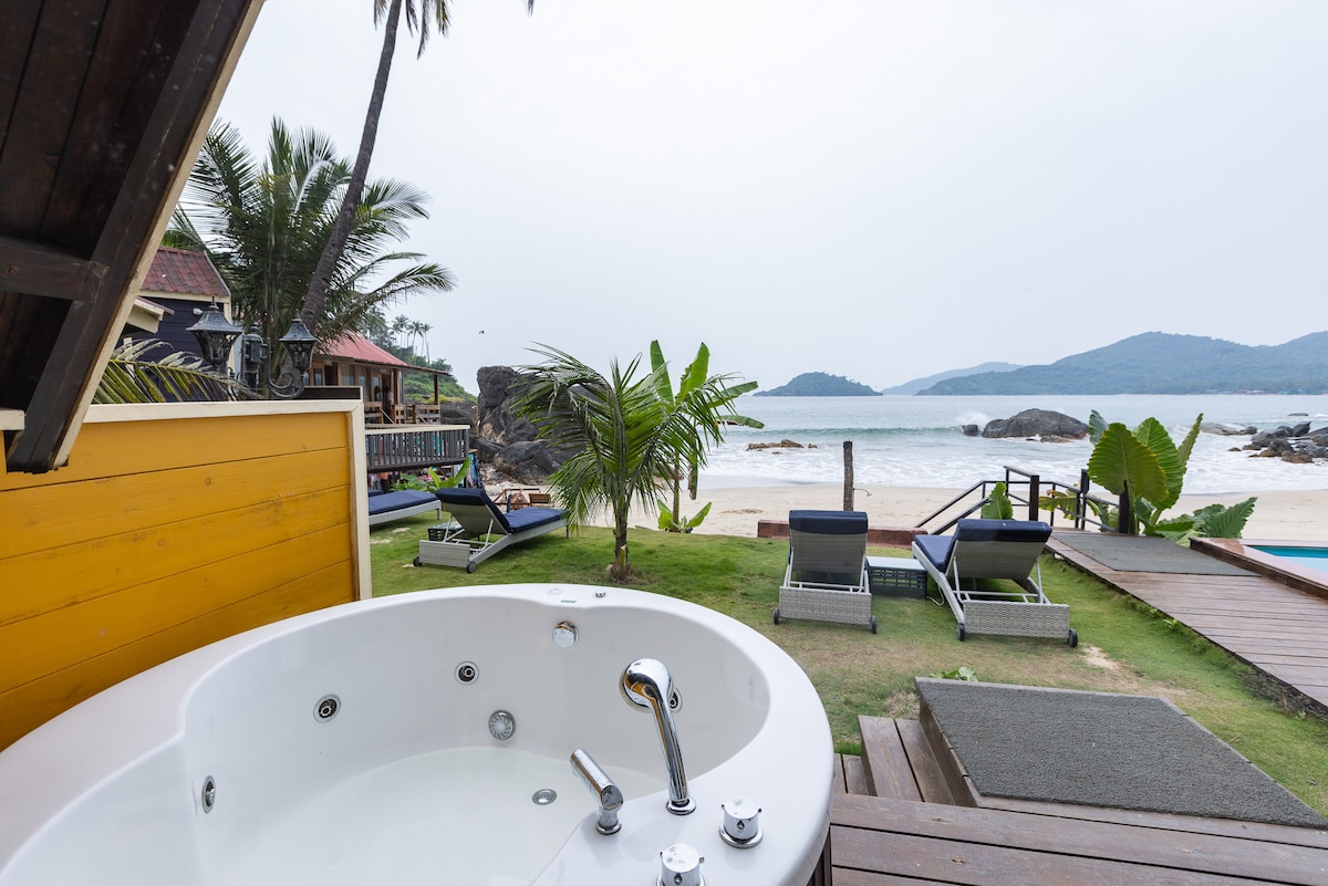 A private outdoor jacuzzi is positioned on a wooden deck, overlooking a sandy beach and the ocean. Sun loungers are arranged on the grassy area beside it, with lush tropical plants and palm trees framing the scene. Rolling hills are visible in the background.