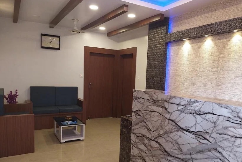A welcoming lobby area features modern furnishings, including a seating arrangement with gray cushions and a dark wooden coffee table. Light streaming from recessed ceiling lights highlights the textured wall and marble reception desk, creating a bright and contemporary first impression.