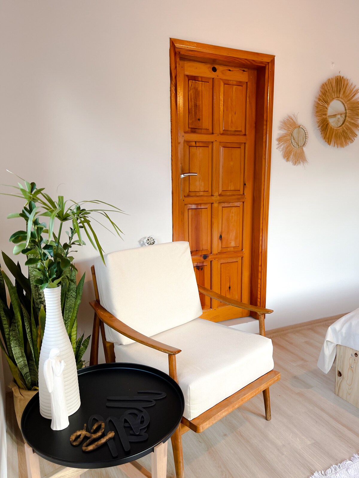 A light-filled corner features a wooden chair beside a dark, circular table. A tall, green plant adds a touch of nature, and a textured white vase is placed on the table. The warm wooden door and round mirrors complement the inviting atmosphere.