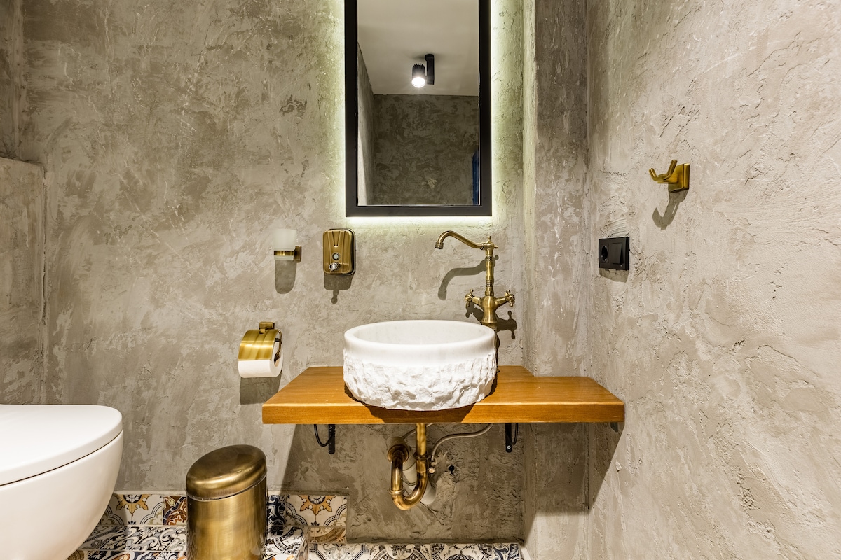 A modern bathroom features a stylish sink set on a wooden countertop against a textured wall. A round mirror is illuminated by soft light, and brass fixtures add a touch of elegance. Decorative tiles enhance the floor design, creating a unique ambiance.