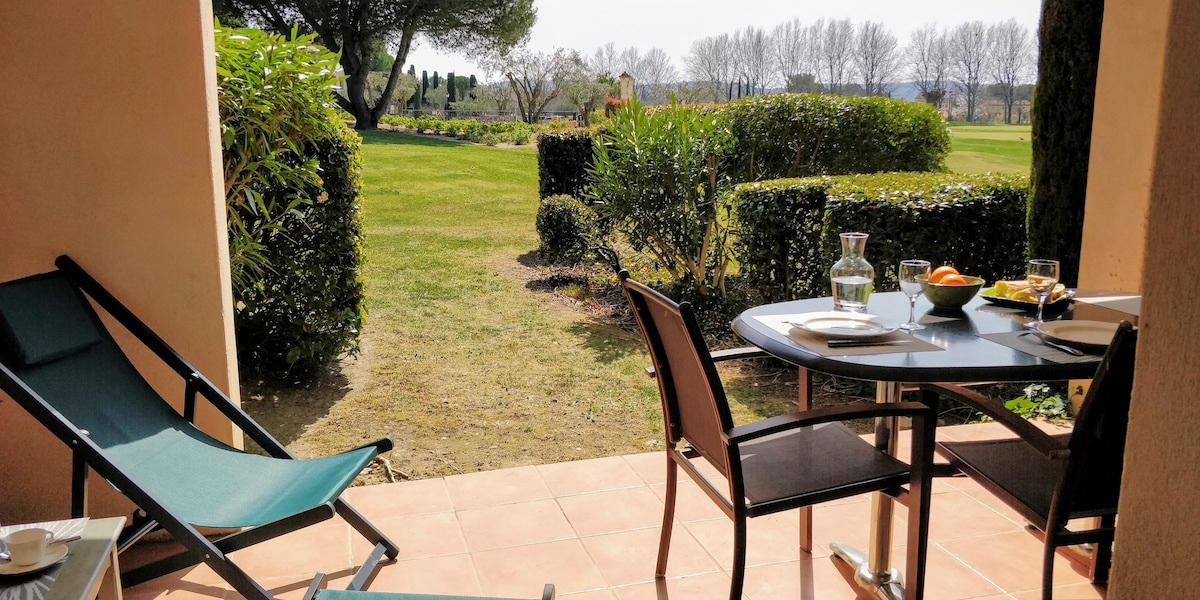 A private patio features a table set for two, accompanied by a pitcher and fresh fruit. Two chairs and a lounge chair are positioned to face the greenery beyond, where manicured hedges and a landscaped lawn create a serene outdoor environment.