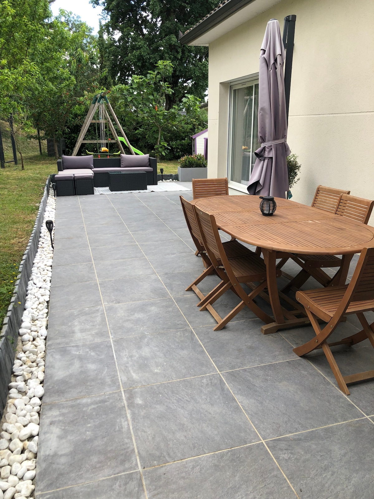 An outdoor patio features a wooden dining table with matching chairs, surrounded by a stone border. A shaded seating area is visible, differentiated by modern furniture. Nearby, lush greenery and a play structure enhance the tranquil atmosphere of the space.