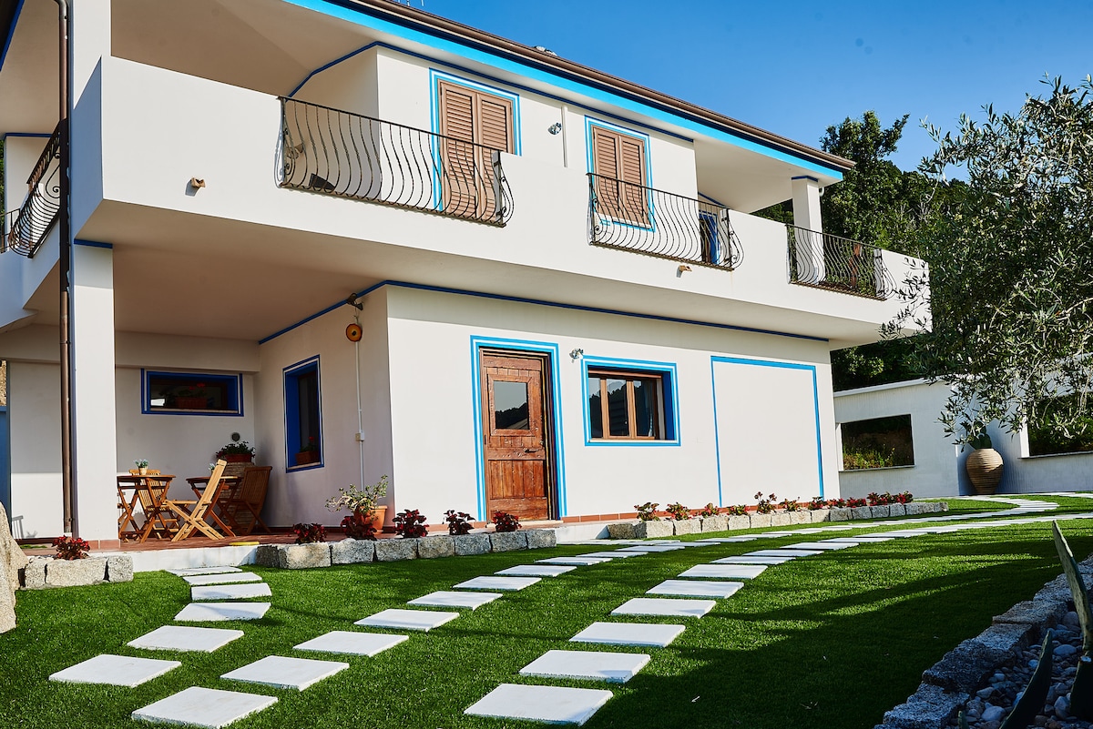 The exterior of the property reveals a two-story structure with a welcoming wooden door. Lush greenery surrounds the home, complemented by a well-maintained pathway of stepping stones leading to a small outdoor seating area with two chairs and a table.