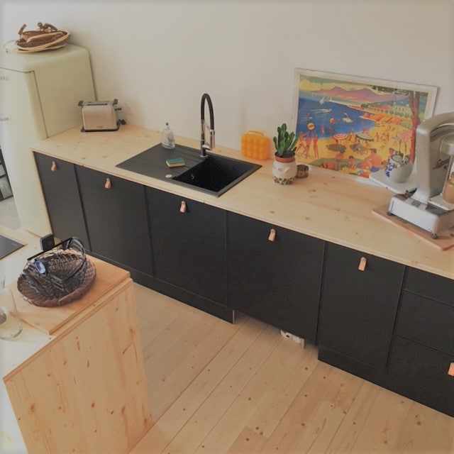 The kitchen features a sleek wooden countertop with a polished black sink and modern faucet. Dark cabinetry provides storage space, complemented by an artwork depicting a beach scene. A toaster and decorative plants add a touch of functionality and warmth to the space.