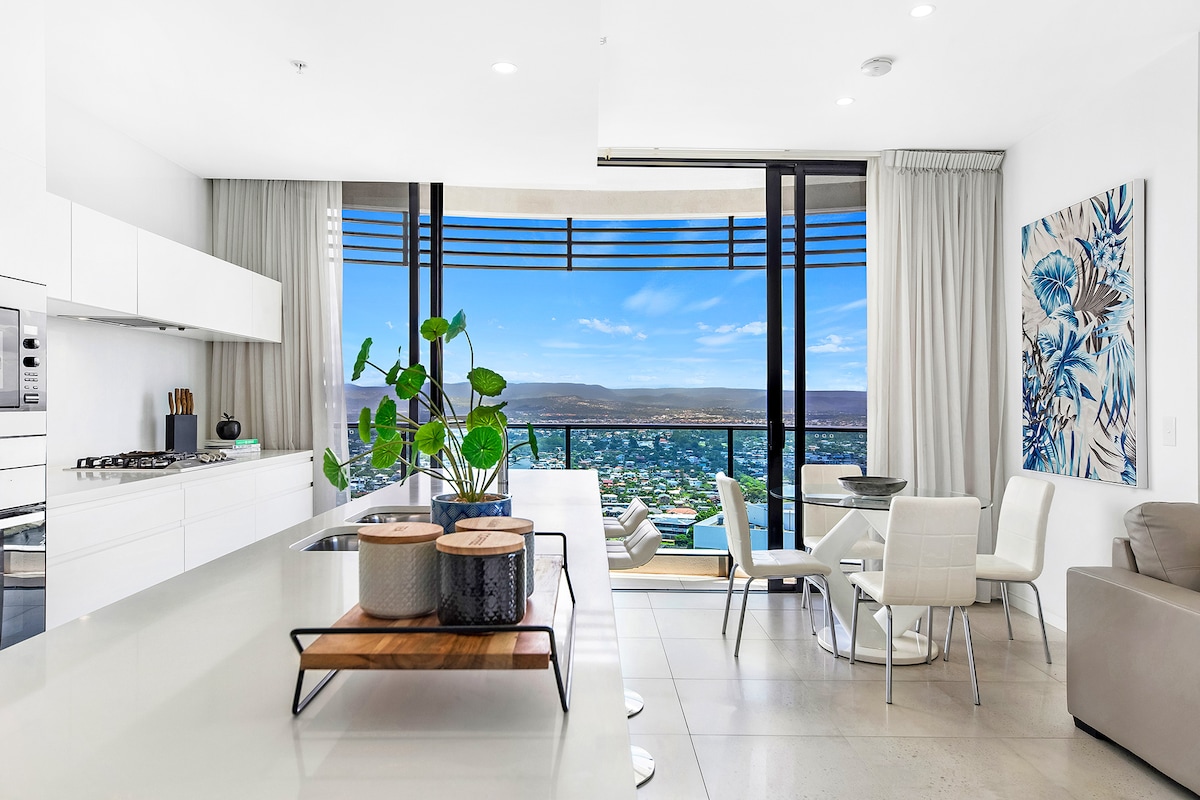 A bright, modern kitchen featuring white cabinetry and stainless steel appliances is showcased alongside a dining area. A large window offers stunning views of the coastline, while elegant art adorns the wall. Neutral-toned furniture complements the spacious, airy atmosphere.