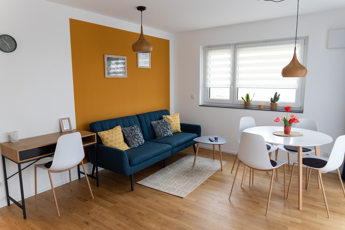 A modern living area features a teal sofa adorned with decorative cushions, complemented by a light-colored area rug. A round dining table with white chairs is surrounded by natural light streaming through large windows. A work desk with a simple design is positioned nearby.