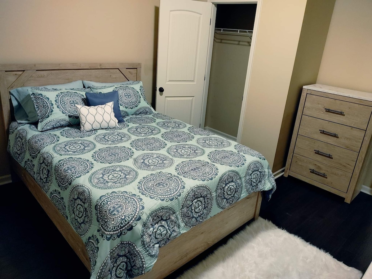 A spacious bedroom features a wooden bed frame with a decorative bedding set in calming blue tones. A small accent pillow complements the bedding. A sturdy dresser is positioned nearby, and a closet with sliding doors is visible in the corner, enhancing storage options.